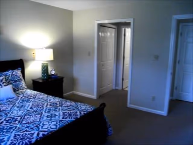 A bedroom with a bed covered in a blue and white patterned bedspread, a nightstand with a lamp and an alarm clock, beige walls, and two open doorways leading to other rooms.