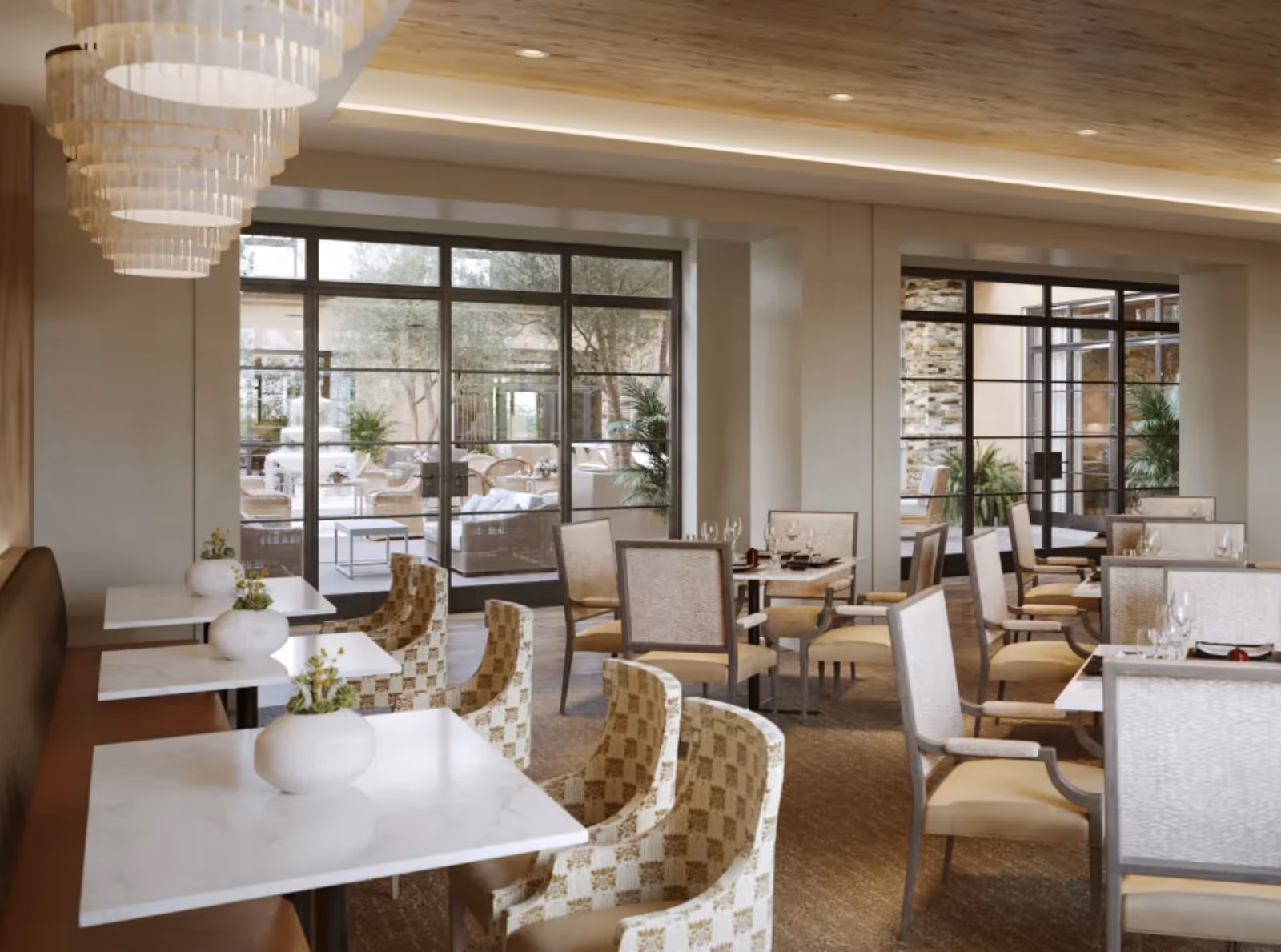 Spacious, elegant dining room with tables and upholstered chairs, chandeliers, and large windows looking out to an outdoor seating area.