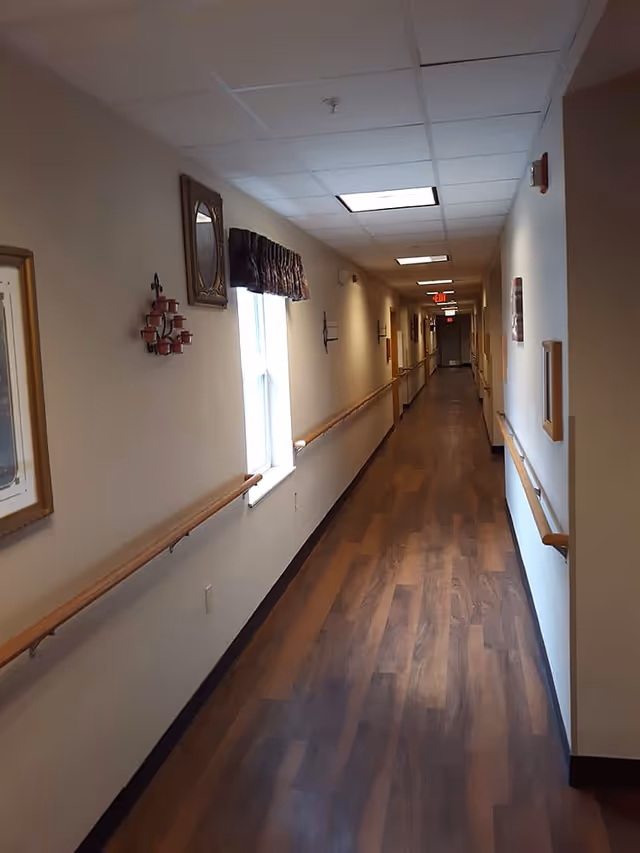 A long, narrow hallway with wooden flooring and handrails on both sides. The walls are decorated with framed pictures, a mirror, and candle holders. There is a window with a valance on the left side, and ceiling lights illuminate the corridor. The hallway appears to be part of a senior living facility.