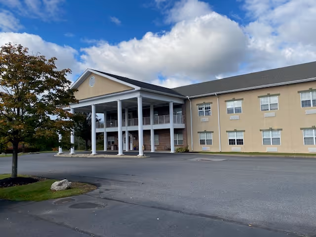 Front exterior of a two-story beige senior living facility with a covered entrance supported by white columns under a partly cloudy sky.