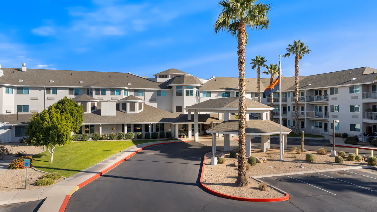 Exterior view of Vista del Rio - A Provincial Senior Living Community, showing a large multi-story building with balconies, a covered entrance, palm trees, and a parking area under a clear blue sky.