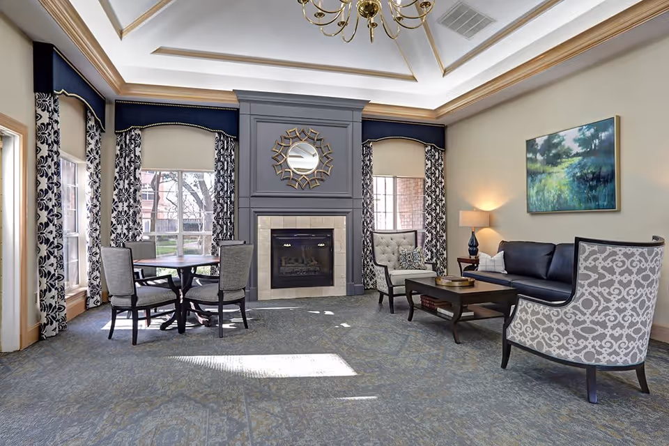 A bright and elegant living room with a fireplace centered on the far wall, flanked by two large windows with patterned curtains. The room features a round table with four chairs on the left, a dark blue sofa, two patterned armchairs, a coffee table, a side table with a lamp, and a landscape painting on the right wall. The ceiling has a tray design with crown molding and a chandelier.