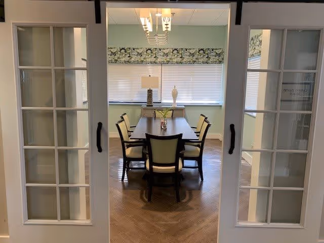 View through white double glass-paneled doors into a private dining room with a rectangular wooden table surrounded by six chairs. The room has light green walls, a window with blinds and a floral valance, a table lamp, and decorative vases on a sideboard against the far wall.