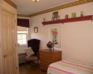 A small bedroom with a single bed covered in a striped blanket, a wooden dresser with a flower vase on top, a cushioned chair with a pillow near a window, and a red wall shelf holding decorative items above a framed floral picture.