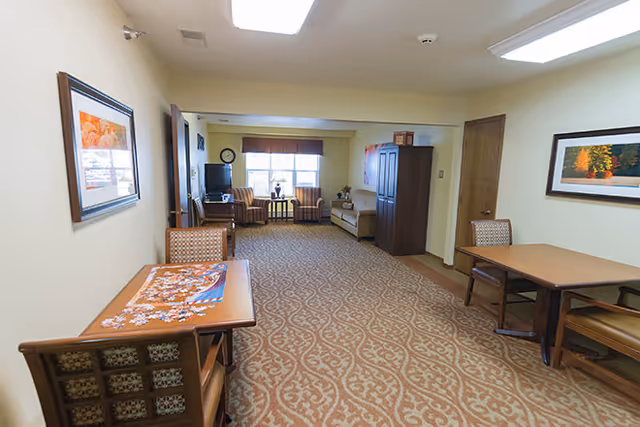 Interior view of a common area in an assisted living facility featuring a carpeted floor with patterned design, two wooden tables with chairs, framed artwork on the walls, and a seating area with armchairs and a TV near a window at the far end.