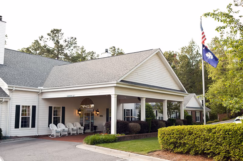 Exterior view of a senior living facility named Golden Bell- Cumberland Hills featuring a covered entrance with white columns, several white rocking chairs, and two flags on a flagpole surrounded by greenery and trees.