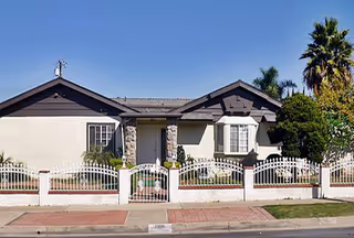 Front exterior view of a single-story residential building with a white fence, stone pillars, and a small garden area with shrubs and palm trees under a clear blue sky.