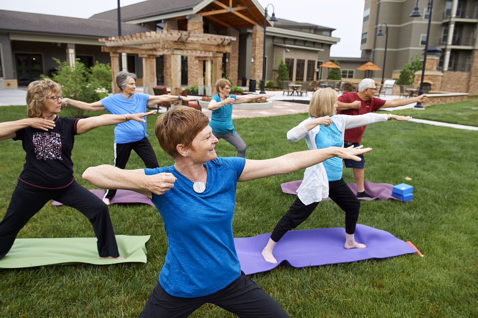 A group of older adults practicing yoga on mats on a lawn in front of a senior living building.