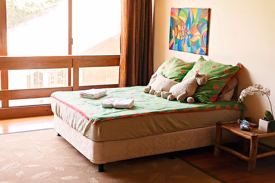 Sunlit bedroom with a double bed dressed in green floral bedding, stuffed animals on the pillows, a bedside table with a plant, and large wooden-framed windows.