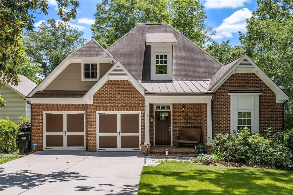 Front exterior view of a single-family brick house with a double garage, a small covered porch with a bench, surrounded by green grass, bushes, and trees under a blue sky with some clouds.