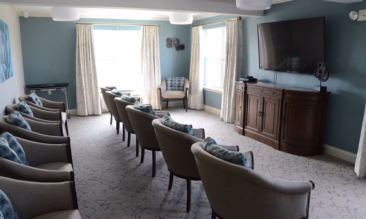 A cozy senior living common room with two rows of beige armchairs, each with a blue patterned cushion, facing a large flat-screen TV mounted on a teal wall above a wooden cabinet. The room has light-colored carpet, two large windows with cream curtains, and a keyboard in the corner.