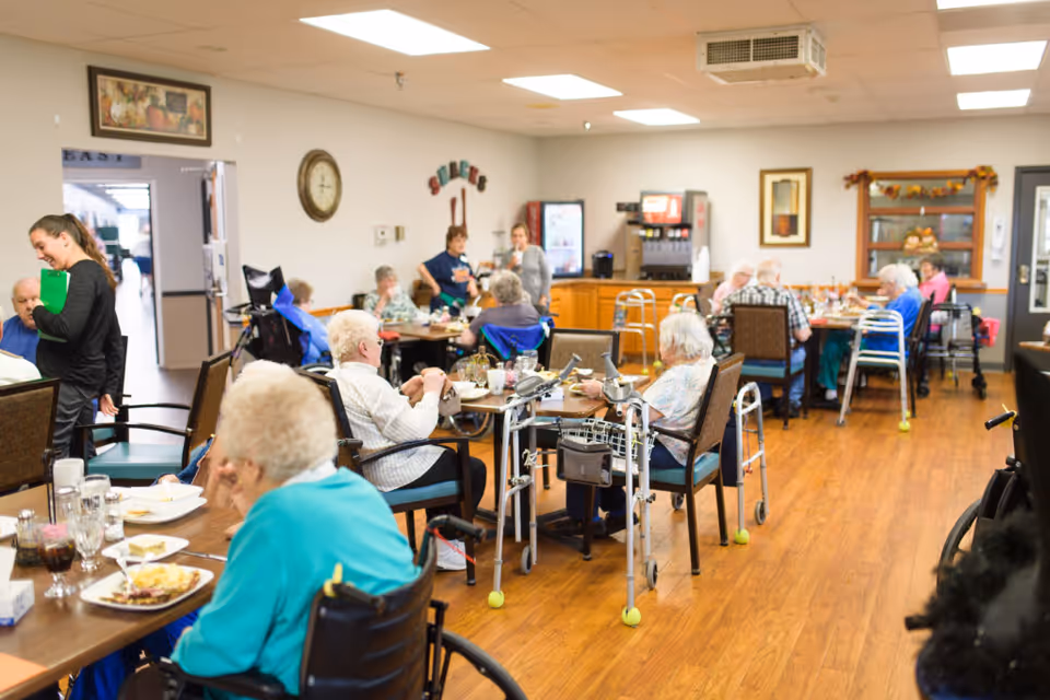 Seniors seated at tables eating and socializing in a communal dining room with staff nearby.