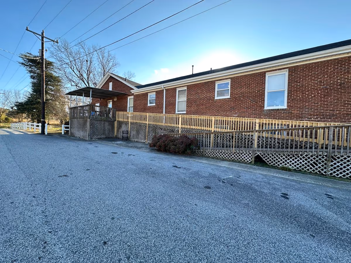 Single-story brick building with a wooden wheelchair ramp and covered porch beside a paved parking area under a clear blue sky.