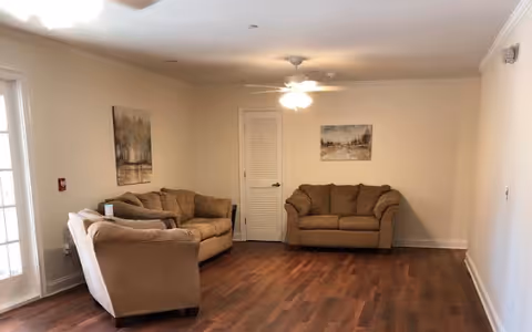 Common sitting room with three beige sofas on wood flooring, a ceiling fan, and framed wall art.