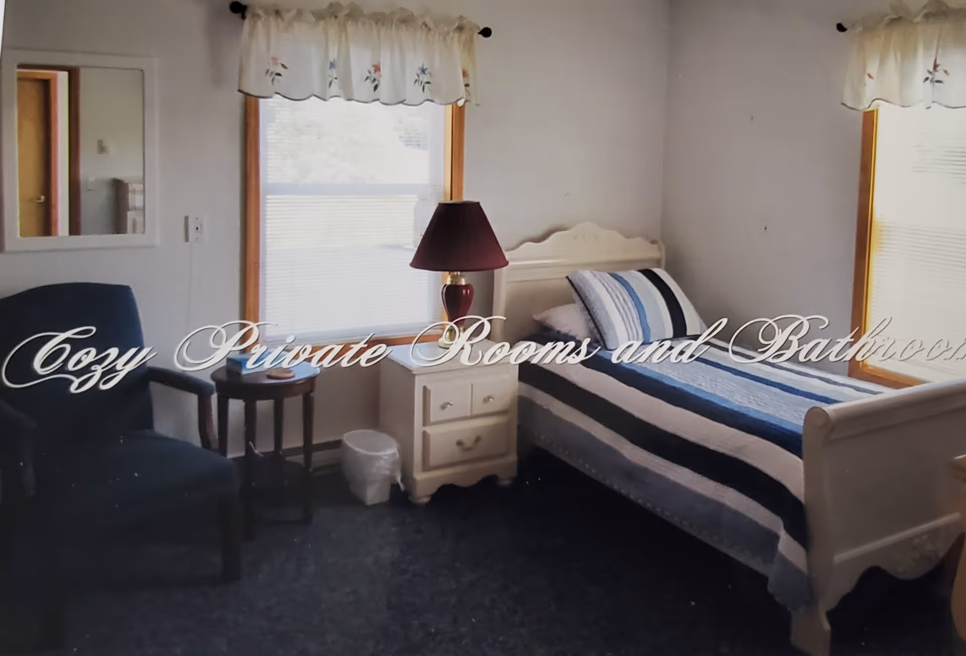 A cozy bedroom with a single bed covered in a striped blue, white, and black quilt. Next to the bed is a white nightstand with a red lamp on it. There is a blue armchair and a small round wooden table beside it. Two windows with floral valances let in natural light, and a mirror hangs on the wall.