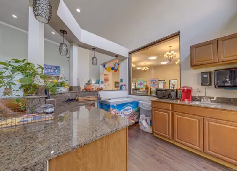 Indoor kitchenette/serving area with granite counters, wooden cabinets, sink, microwave and a Keurig with a pass-through window to a decorated activity room.