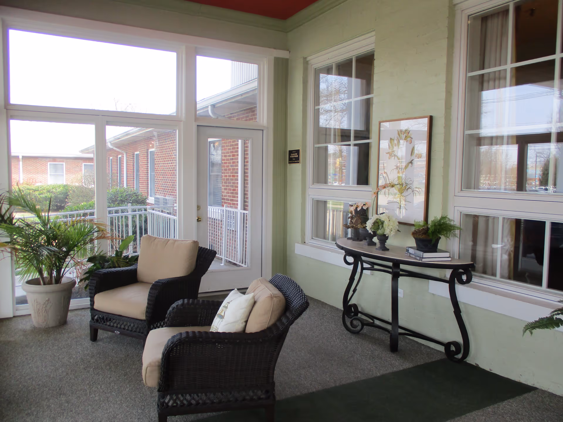 A cozy indoor seating area with two cushioned wicker chairs, a small table with decorative plants and books, large windows, and a glass door leading outside. There are potted plants near the windows and a framed artwork on the wall.