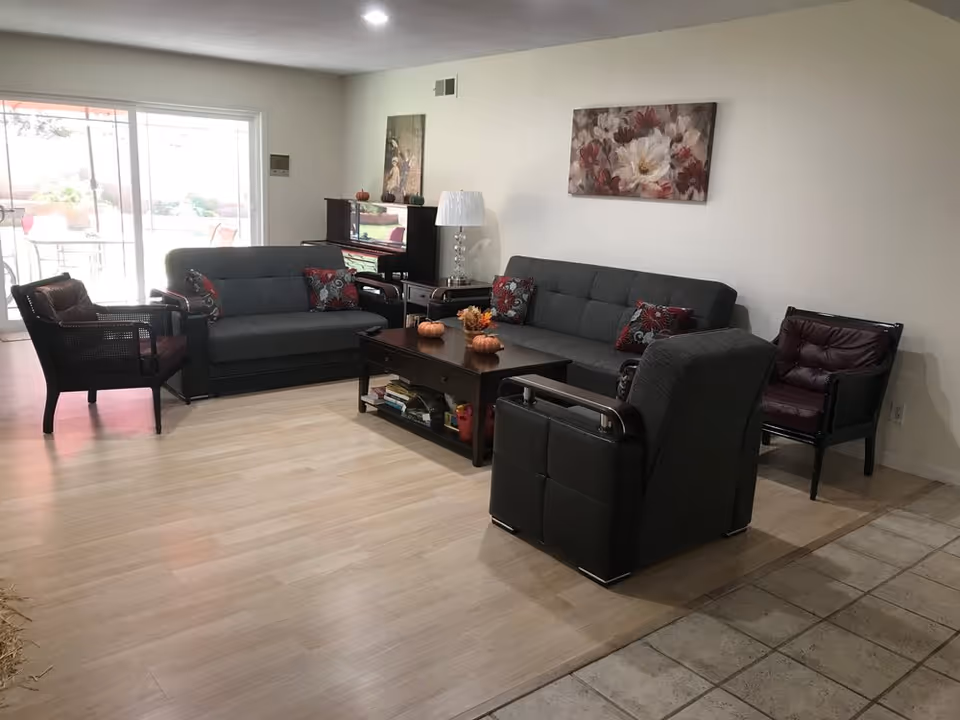 A cozy living room with two gray sofas, two dark armchairs with maroon cushions, a wooden coffee table with small decorative pumpkins and flowers, a side table with a lamp, a piano against the wall, and floral artwork hanging above the sofa. Large sliding glass doors lead to an outdoor patio area.