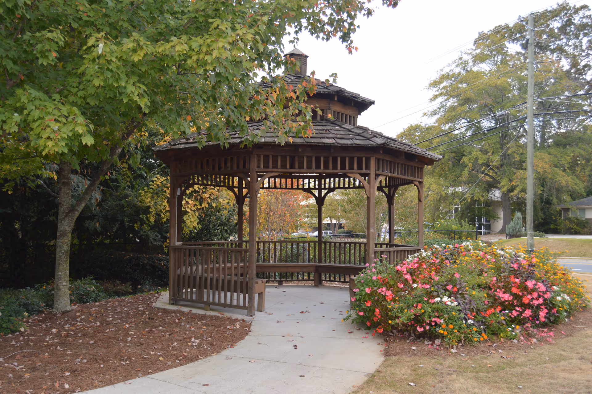 A wooden gazebo with a shingled roof situated in a garden area. The gazebo is surrounded by trees with green and autumn-colored leaves, and a flower bed with vibrant pink, orange, yellow, and white flowers. A concrete pathway leads to the gazebo, and there are power lines and a road visible in the background.