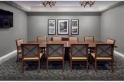 A formal meeting or dining room with a long wooden table surrounded by ten wooden chairs with cushioned seats. The walls are painted gray and decorated with three black and white framed pictures. The floor is carpeted with a patterned design, and two chandeliers hang from the ceiling.
