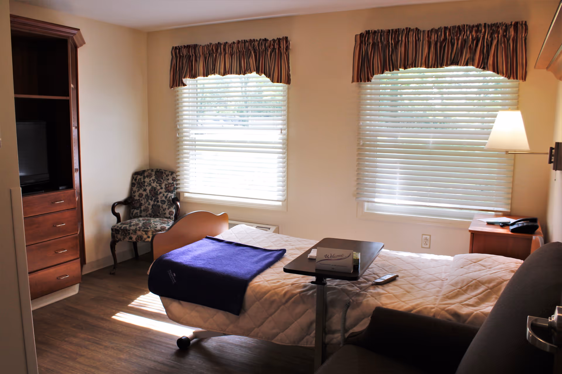 Furnished bedroom with a single bed, overbed table, armchair, TV cabinet, and two windows with blinds.