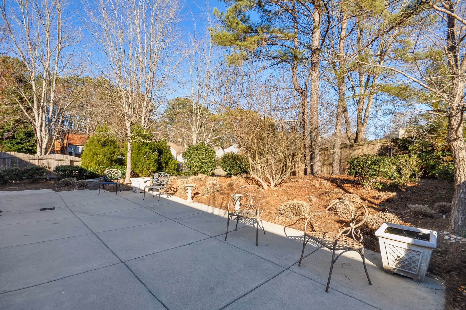 Outdoor patio with metal chairs and planters beside landscaped beds and trees under a clear blue sky.