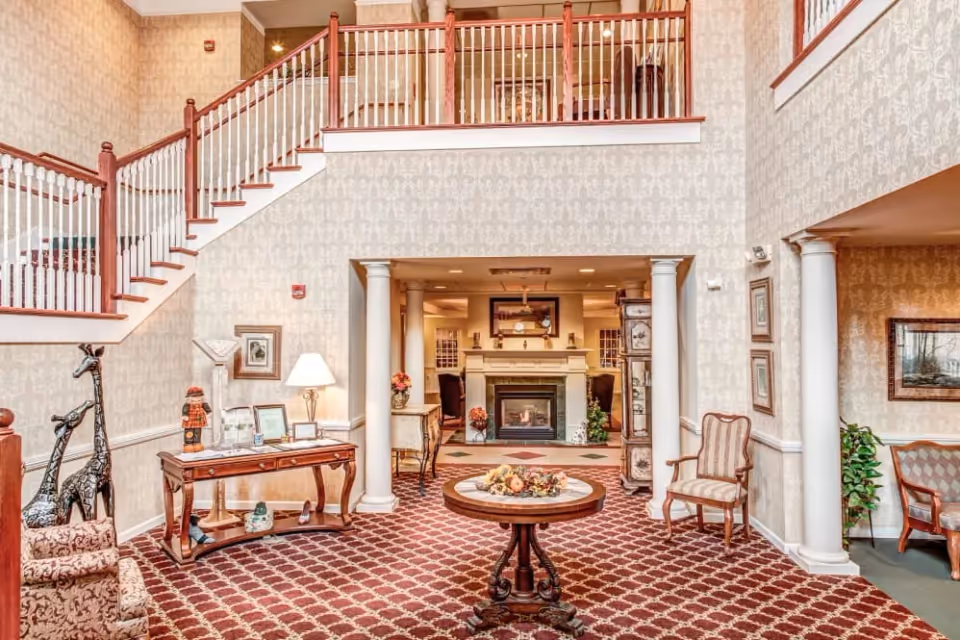 A spacious and elegantly decorated senior living facility common area with a high ceiling and a staircase with wooden railings. The room features a patterned red carpet, a round wooden table with a floral centerpiece, two armchairs, decorative giraffe sculptures, framed artwork on the walls, and a fireplace in the background flanked by columns.