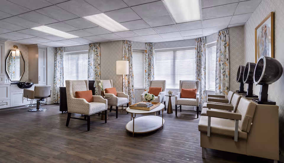 Bright, comfortable salon-style lounge with armchairs, a coffee table, hair-dryer stations and salon chairs in front of bay windows.
