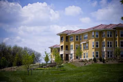 Exterior view of a yellow senior living facility on a grassy slope under a cloudy sky.