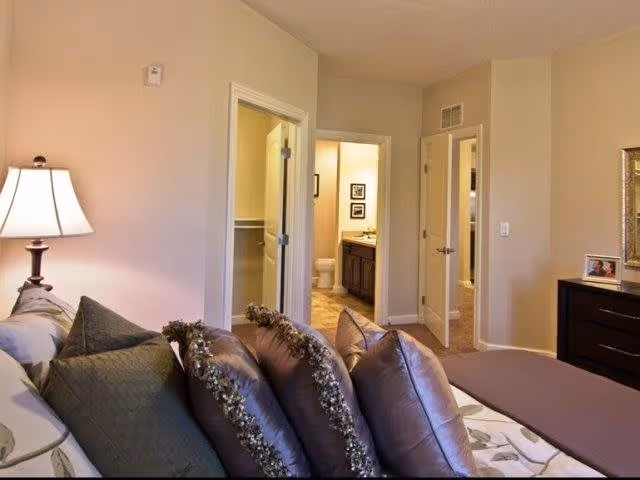 A cozy bedroom with a bed featuring multiple pillows in shades of purple and green. To the left, there is a lit table lamp on a nightstand. In the background, an open door reveals a bathroom with a toilet and vanity, and another door leads to a closet or another room. A dresser with framed photos is visible on the right side of the room.