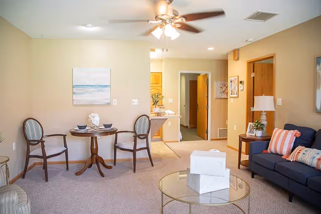 Well-lit living room with a small round dining table and chairs, a navy sofa with pillows, glass coffee table, and an open doorway to a hallway.
