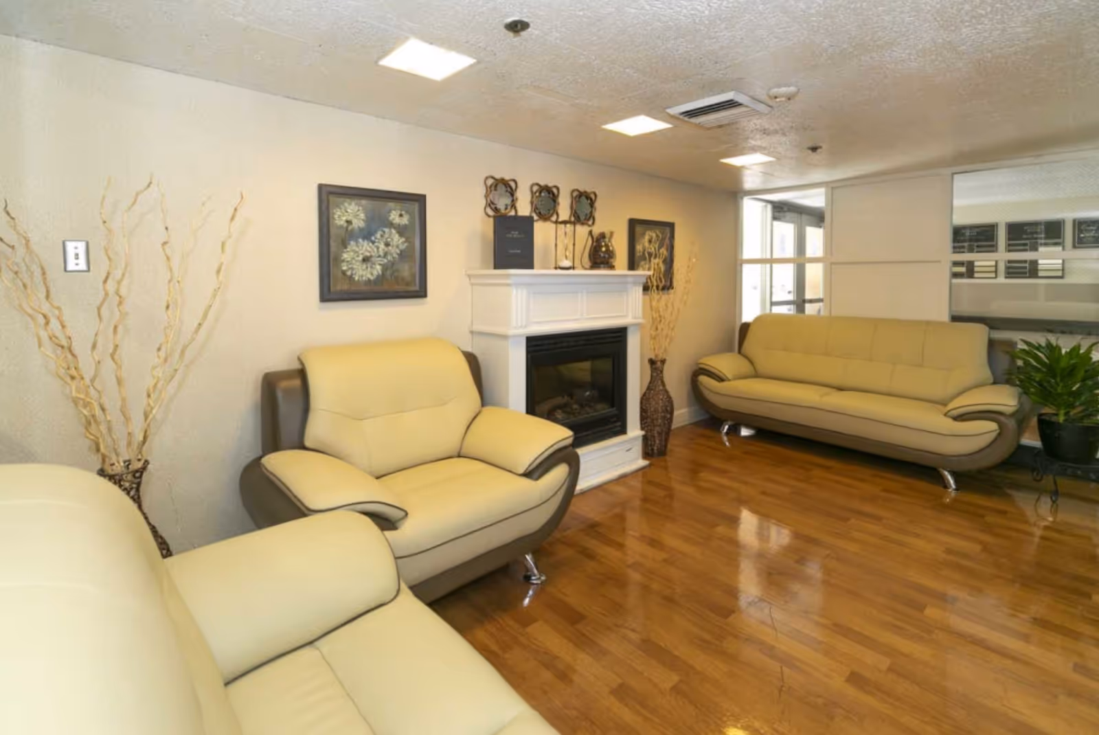 A cozy living room area with three beige and brown leather sofas arranged around a white electric fireplace. The room has wooden flooring, light-colored walls, and decorative elements including framed floral artwork and tall vases with dried branches. There is a large window or glass partition on one side letting in natural light.