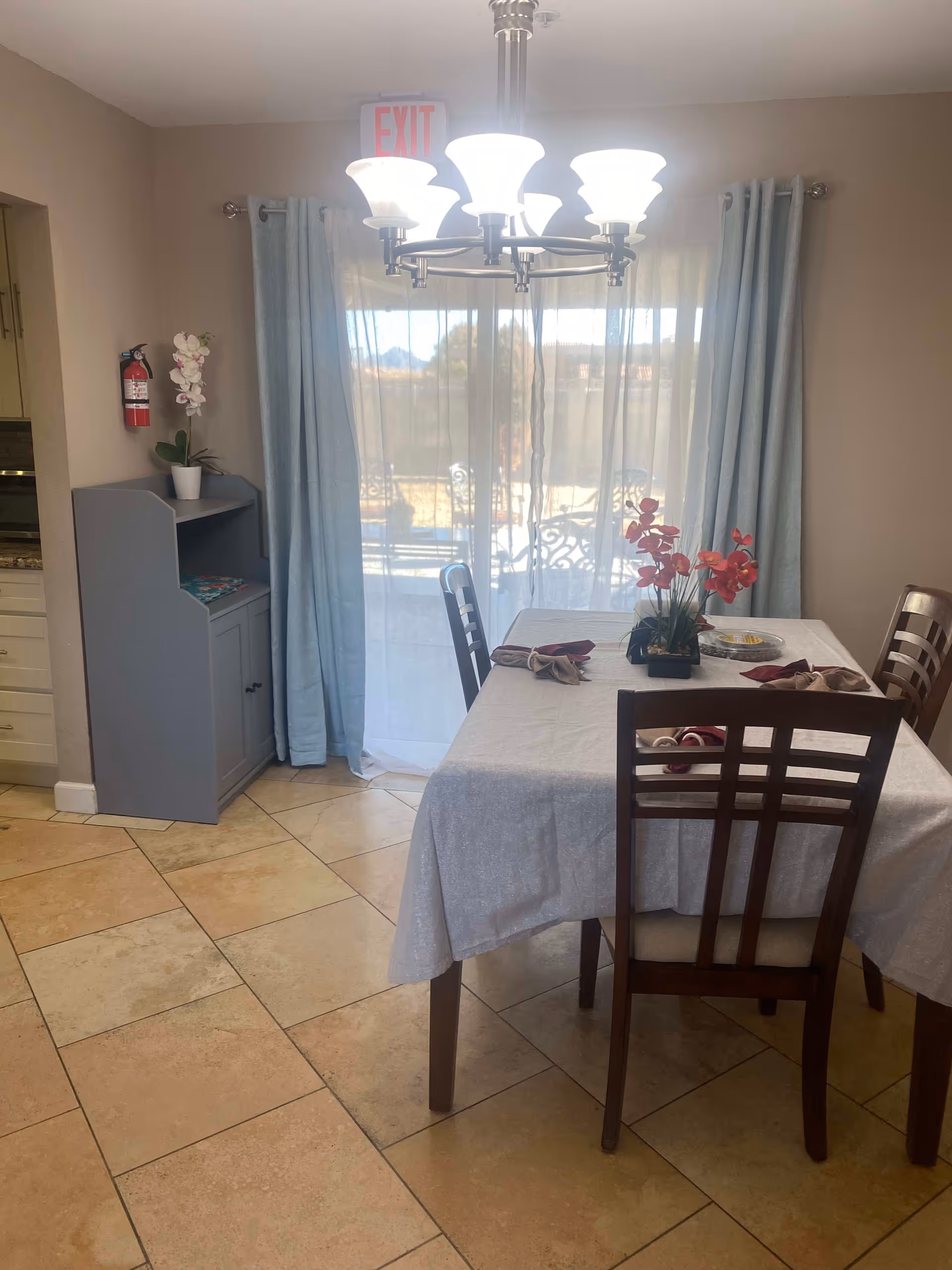 A dining area with a rectangular table covered with a white tablecloth and four wooden chairs. The table is set with folded napkins and a centerpiece of red flowers. Behind the table is a large window with sheer white curtains and blue drapes, allowing natural light to enter. A chandelier with multiple lights hangs above the table. To the left, there is a small gray cabinet with a potted orchid on top and a fire extinguisher mounted on the wall. The floor is tiled with large beige tiles.