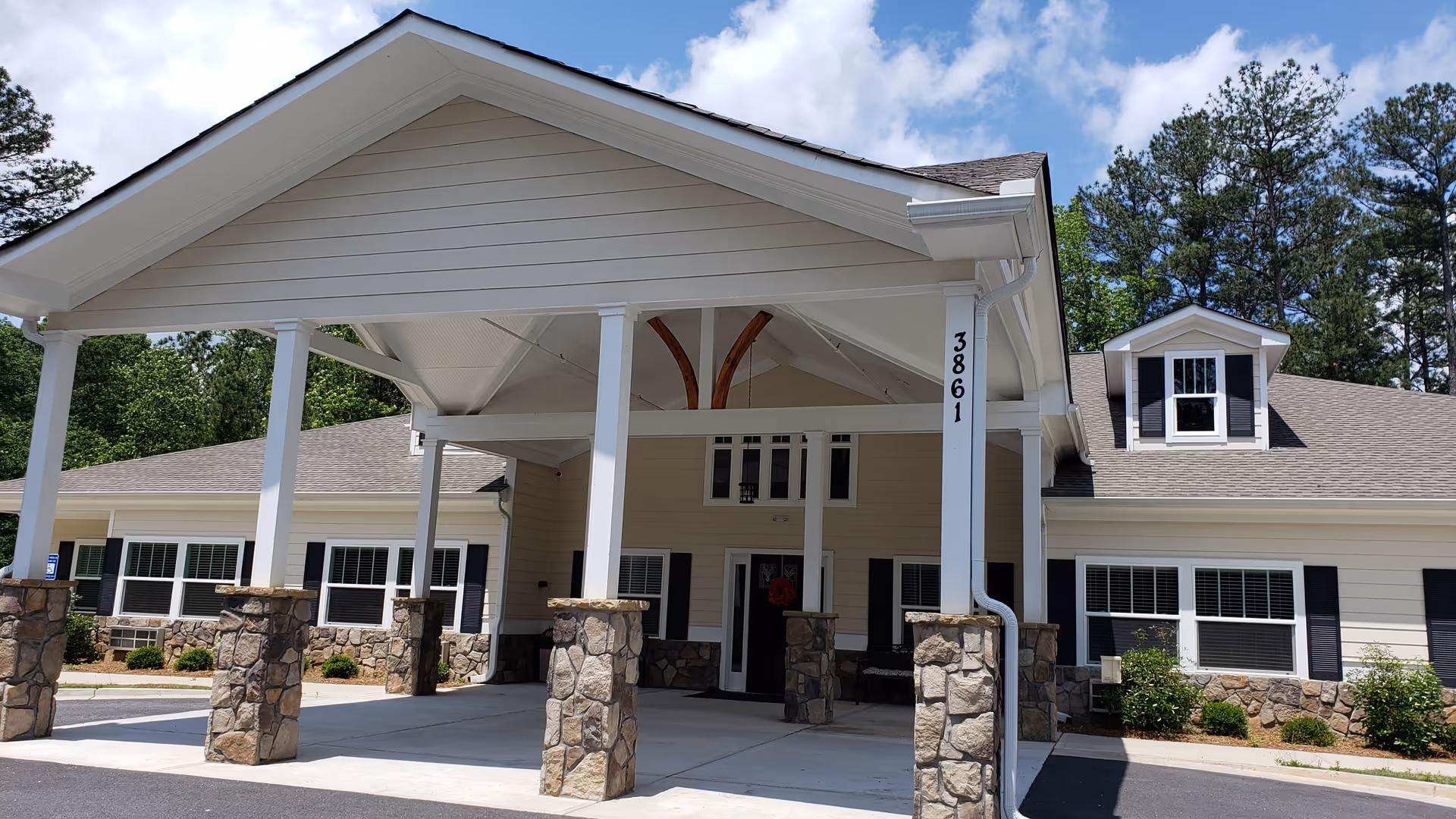 Front exterior view of a senior care home building with a large covered entrance supported by white columns with stone bases. The building has beige siding, black shutters, multiple windows, and a gray shingled roof. The address number 3861 is visible on one of the columns. Trees and a partly cloudy sky are in the background.
