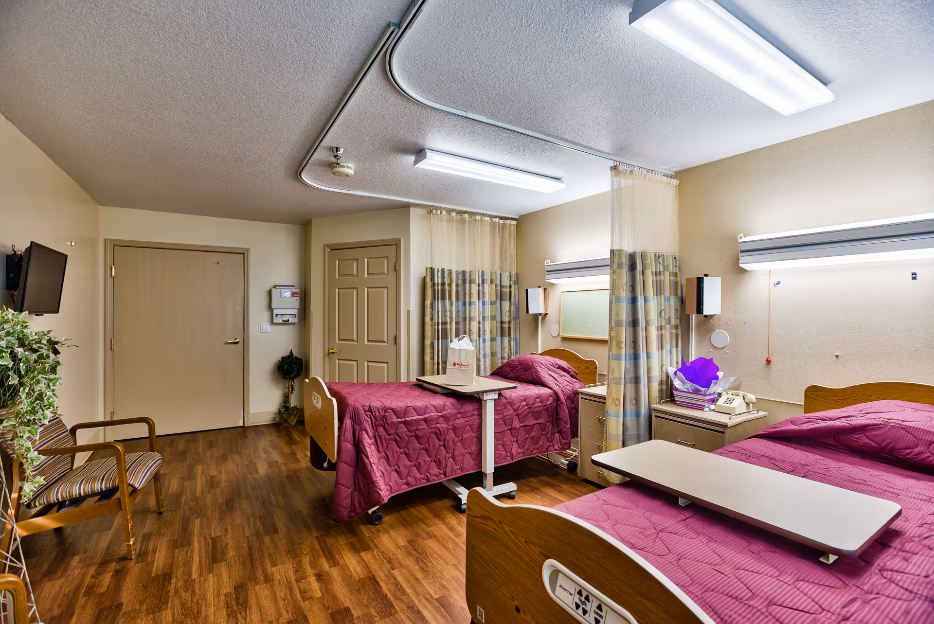 A shared patient room in a senior living facility with two hospital-style beds covered in maroon quilts, each with an overbed table. The room has wood flooring, beige walls, a wall-mounted TV, a chair, and privacy curtains between the beds. There are overhead fluorescent lights and a door at the back of the room.