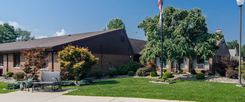 Exterior view of Bridgeport Healthcare Center showing a single-story brick building with a sloped roof, surrounded by well-maintained landscaping including trees, shrubs, and a green lawn. There is a bench on a concrete pathway and an American flag on a flagpole near the building.
