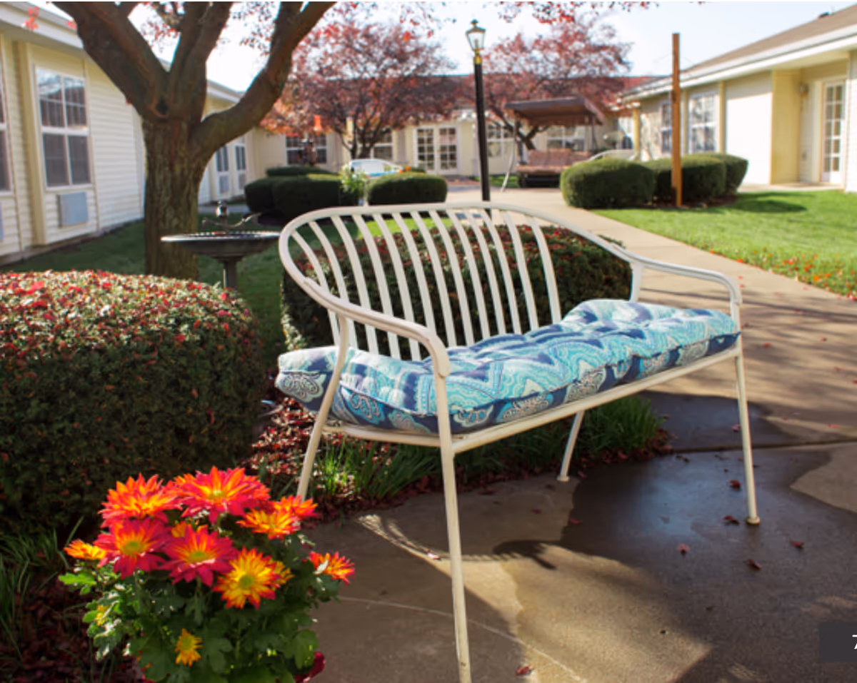 Outdoor courtyard area at Honeysuckle Senior Living featuring a white metal bench with a blue patterned cushion, vibrant red and yellow flowers in the foreground, trimmed bushes, a tree, and a paved walkway surrounded by single-story buildings.