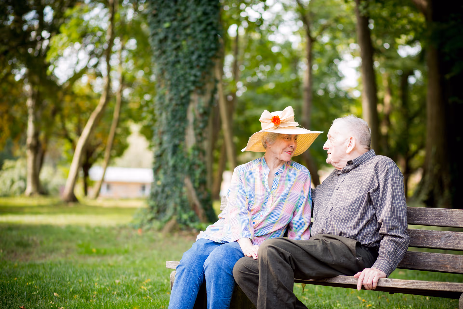 An elderly woman wearing a large sunhat with a flower and a plaid shirt sits on a wooden bench outdoors, smiling and looking at an elderly man in a checkered shirt and dark pants who is sitting next to her. They are surrounded by green grass and trees in a park-like setting.