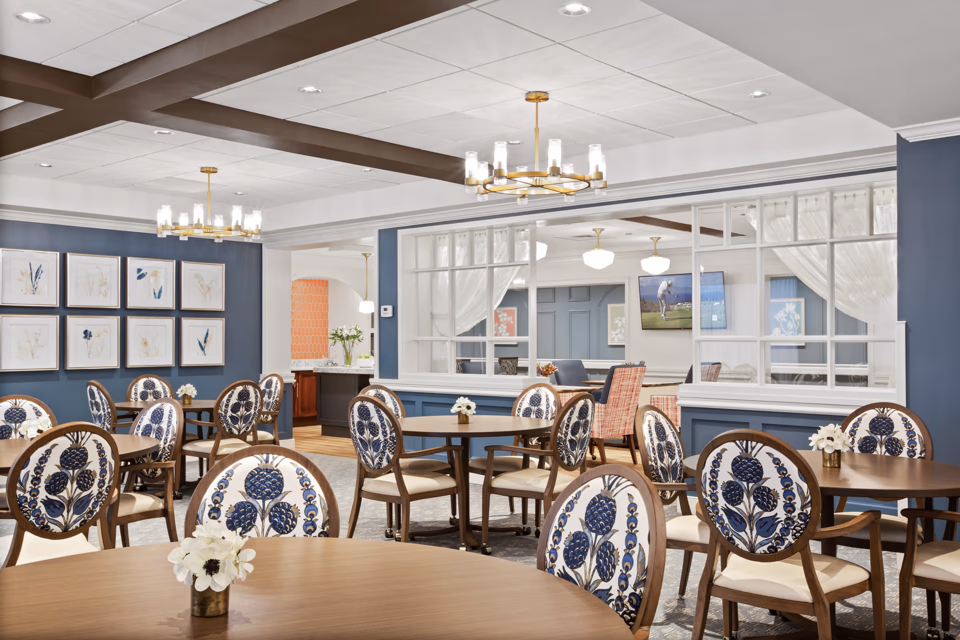 A bright and elegant dining room with round wooden tables and chairs featuring blue floral upholstery. The walls are painted blue with framed botanical artwork. Two modern chandeliers hang from the ceiling. A large window with white curtains separates this room from a cozy sitting area with armchairs and a wall-mounted TV.