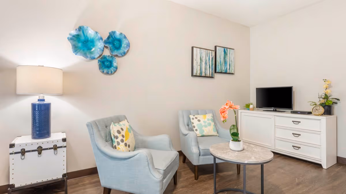 A cozy living room area with two light blue armchairs featuring patterned cushions, a small round coffee table with a potted orchid, a white sideboard with a flat-screen TV, and decorative wall art including blue floral sculptures and framed paintings. A blue table lamp sits on a white chest next to one of the chairs.