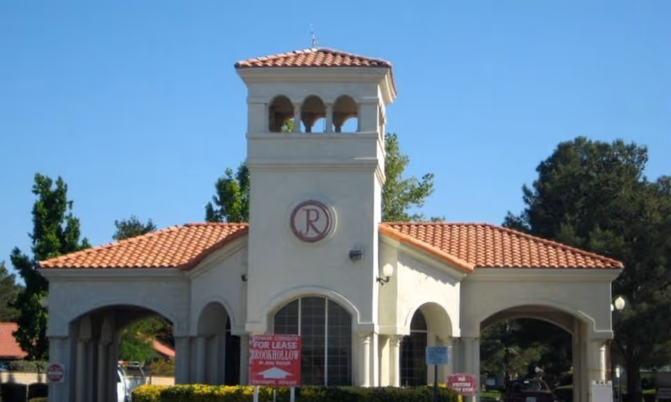 Mediterranean-style entrance building with a tower and red tile roof, a circular 'R' emblem, and 'For Lease' signs in front.