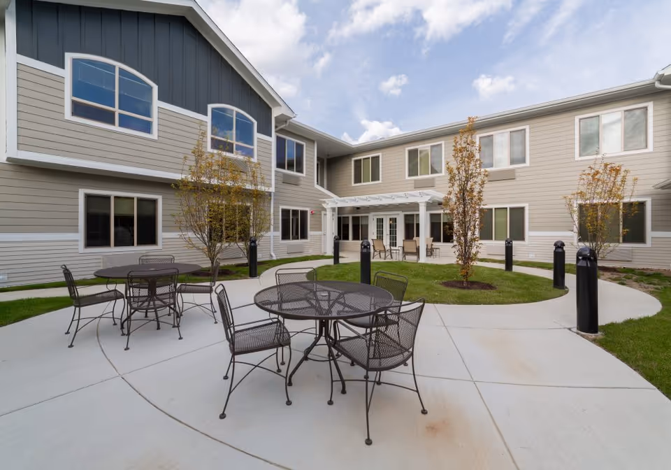 Courtyard patio with metal tables and chairs and small trees in front of a two-story senior living building under a partly cloudy sky.