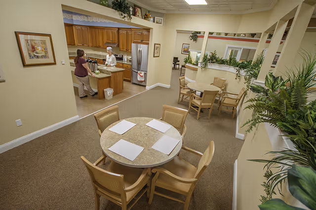 Interior view of a senior living facility dining area with round tables and chairs. Two women are conversing near a kitchen area with wooden cabinets and a stainless steel refrigerator. The room is decorated with plants and framed pictures on the walls.
