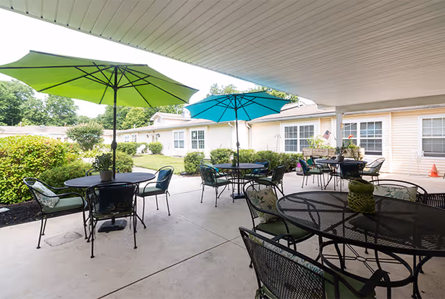 Outdoor patio area at Granville Place with multiple round metal tables and chairs, some tables shaded by large green and blue umbrellas. The patio is adjacent to a single-story building with beige siding and white-framed windows, surrounded by green bushes and trees.
