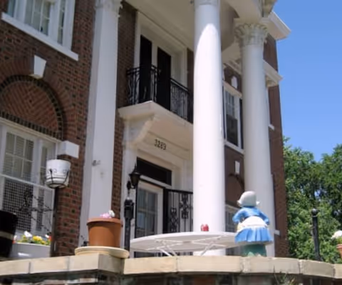 Front exterior view of a brick building with large white columns and a balcony above the entrance. There is a white round table with a small statue of a girl in a blue dress on the patio in front of the building. The building number 3249 is visible above the door.