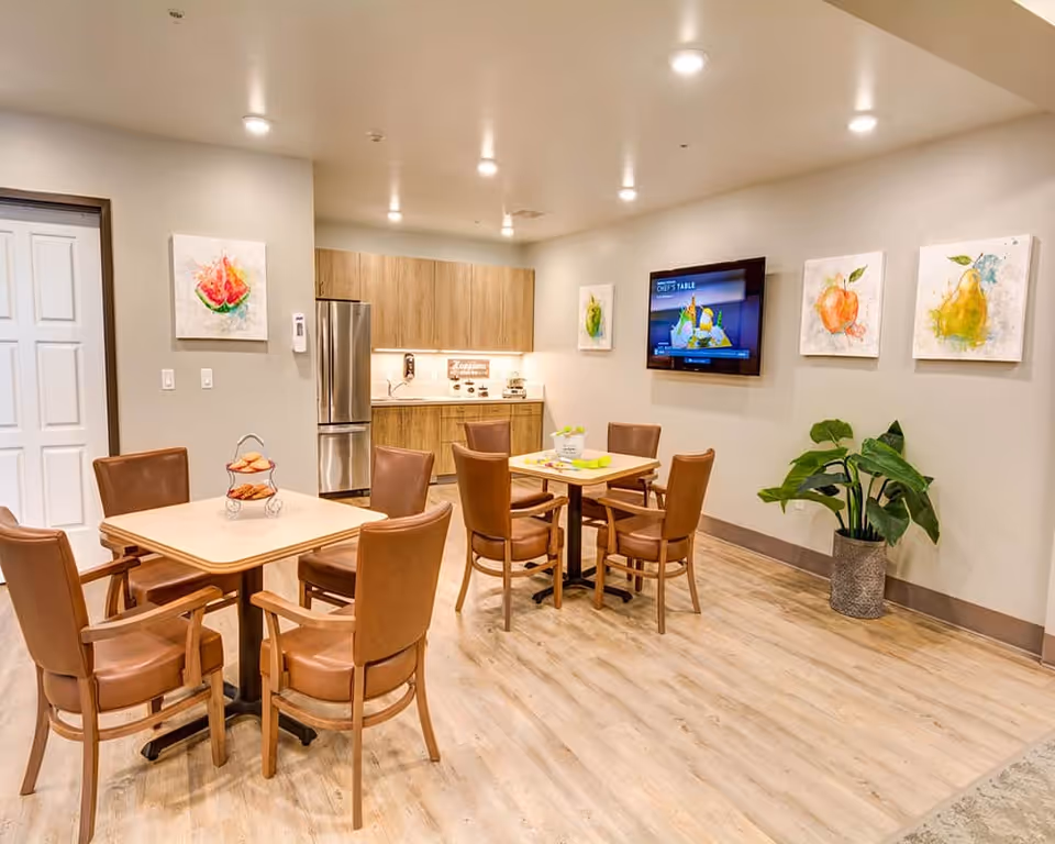 Bright communal dining area with multiple tables and chairs, a small kitchenette, wall-mounted TV, artwork, and a potted plant.
