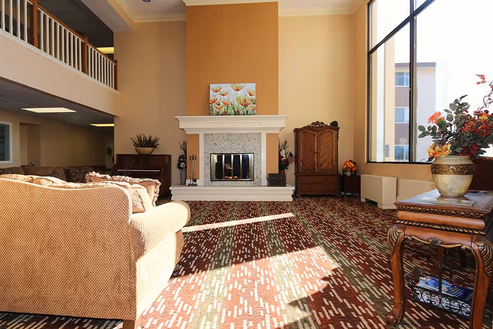 Sunlit communal living room with a fireplace, sofa, patterned carpet and large windows.