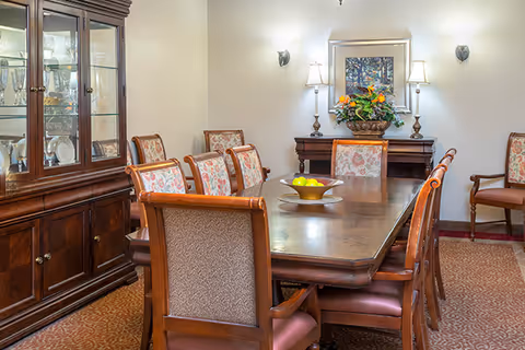 A formal dining room with a long wooden table surrounded by eight upholstered chairs. A wooden china cabinet with glass doors displaying glassware is on the left. On the far wall, there is a sideboard with a floral arrangement and two lamps, and a framed picture above it. The room has soft lighting and a patterned carpet.