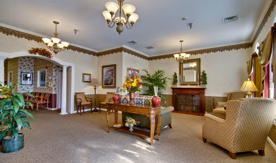 A cozy living room area in a senior living facility with beige patterned wallpaper and carpet. The room features a wooden table with flower arrangements, a green sofa with patterned cushions, two upholstered armchairs, a decorative fireplace with a mirror above it, and several plants. There are two chandeliers hanging from the ceiling and a window with curtains letting in natural light. An archway leads to another room with wooden chairs and floral wallpaper.