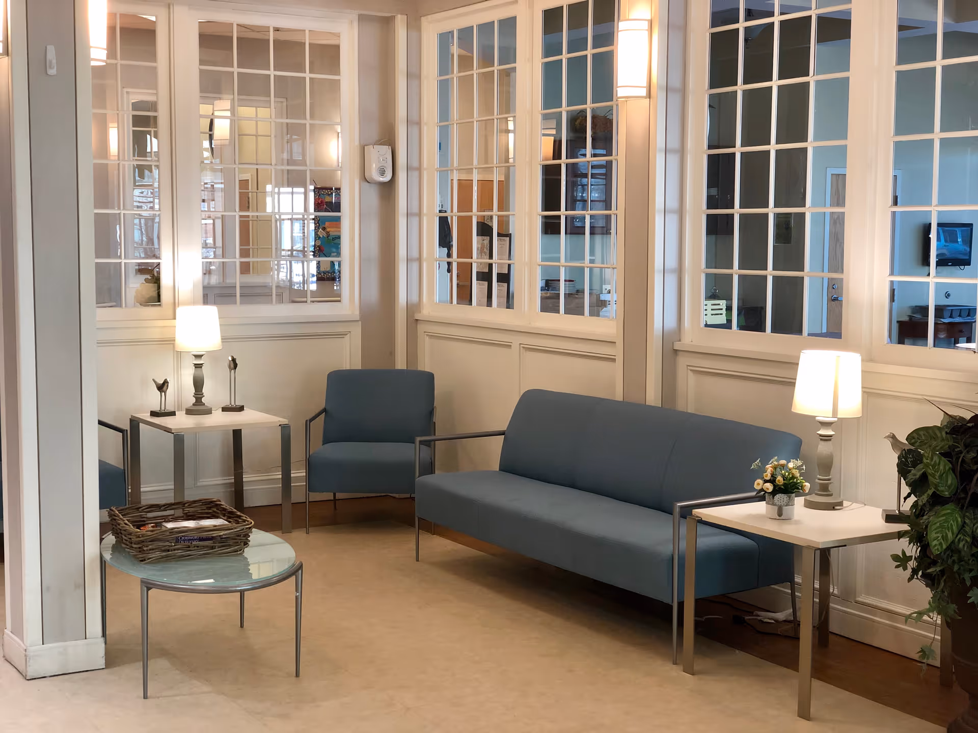 A bright interior seating area with a blue sofa and chairs, side tables with lamps, a glass coffee table and paned interior windows.